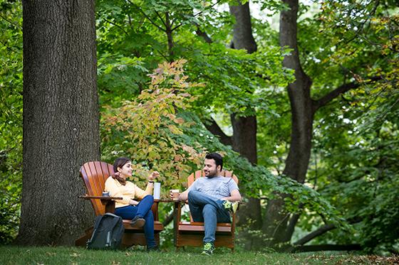 图为两名波胆网站的学生坐在外面绿草地上高大的橡树下的阿迪朗达克椅子上喝咖啡. 